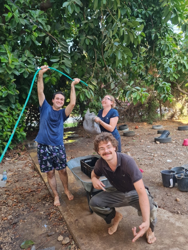 Eitan Cassway, Asya Orlov, and Arkady Boitsov outside with a water hose, wheel barrow, planters on a wooden plank, with trees in the background.